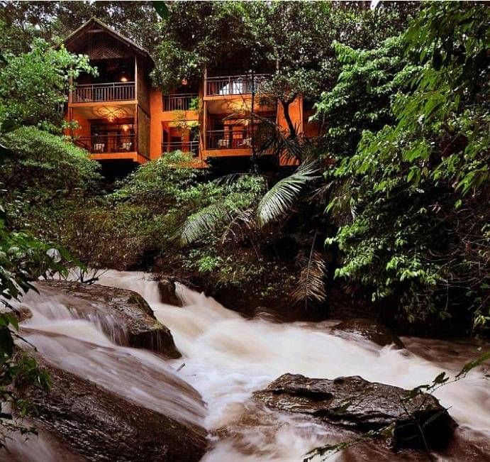 A view of cottages nestled in greenery, from the rocks near the water stream | Vythiri Resort, Wayanad